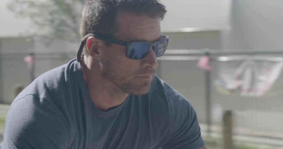 A man wearing sunglasses concentrates as he rides his bike past a park and buildings
