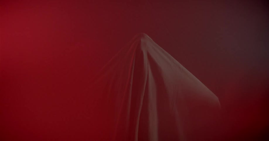 A person stands in smoky red light wearing a sheet like they were a ghost