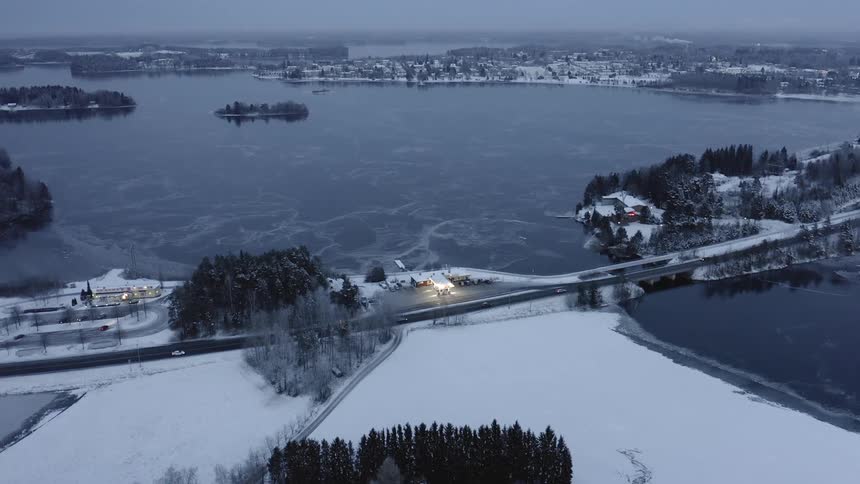 Cars drive along a road that passes a large body of water as snow coats the ground nearby