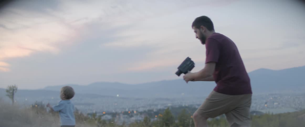 A dad uses a camcorder to film his toddler on a hillside trail at sunset
