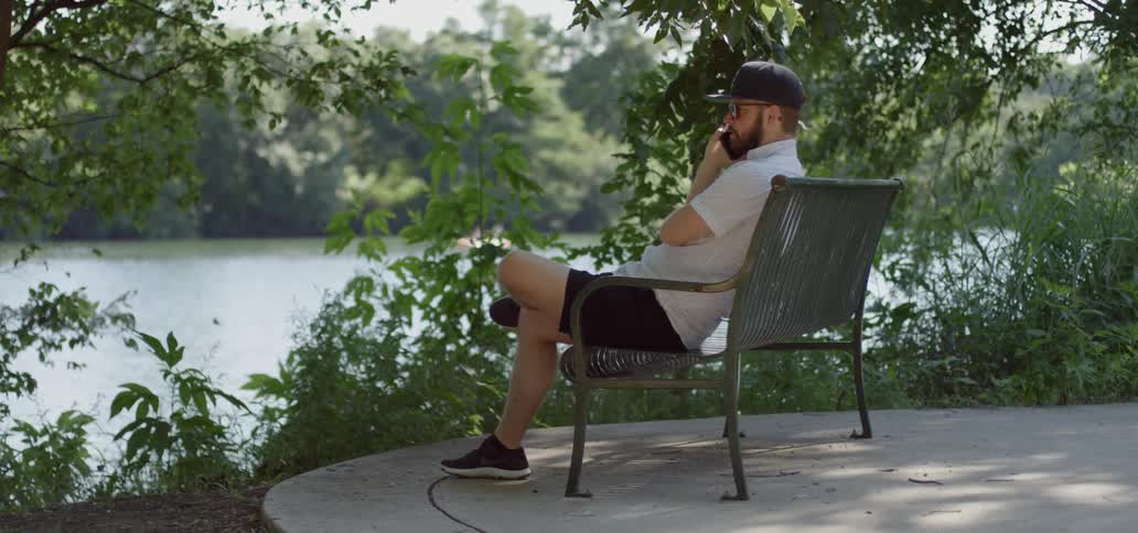 A man sits on a bench by a river and talks on a phone