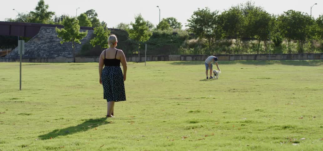 A man and a white dog are playing at a park while a female in a dress is slowly approaching them