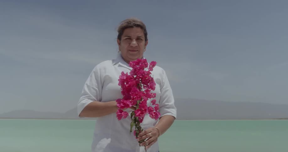 A woman stands in front of the water and holds flowers