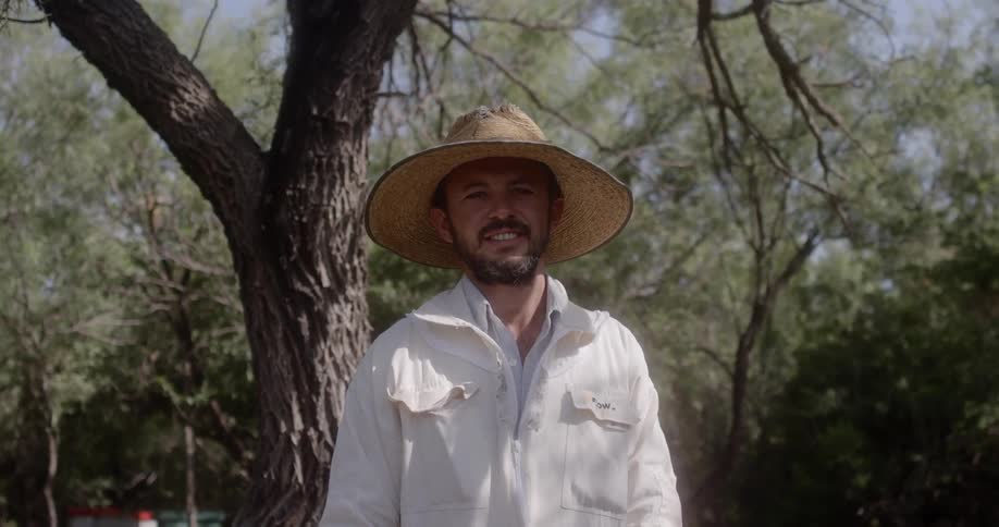 A smiling man wearing a straw hat and white coveralls stands near trees