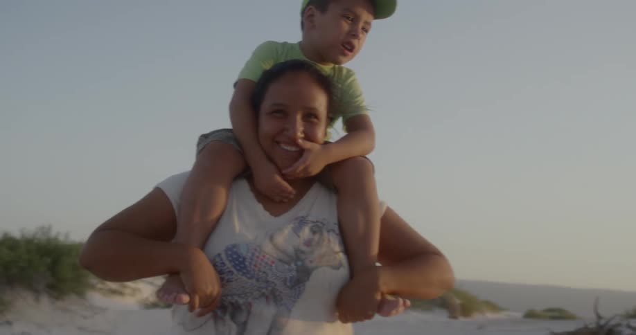 A woman stands outside on a beach and smiles as a young boy sits on her shoulders