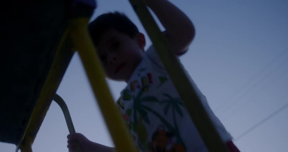 A young boy climbs up a ladder leading to a playground slide at sunset