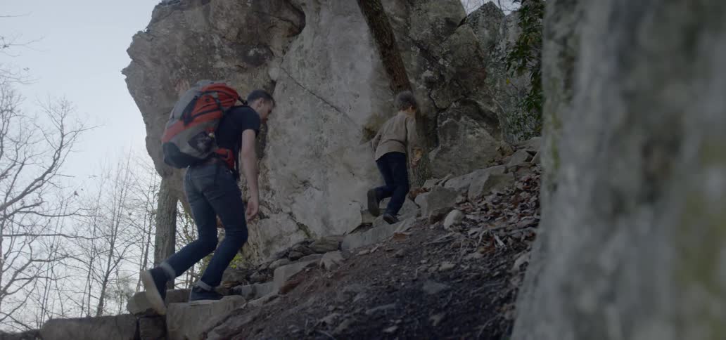 A dad wearing a large backpack climbs up a rocky hill behind his young son