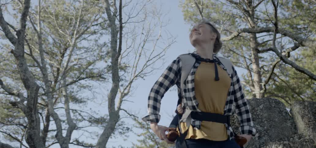 Mother carries toddler on back in a hiking carrier and looks up to the sky through the trees in the woods feeling the wind on her face