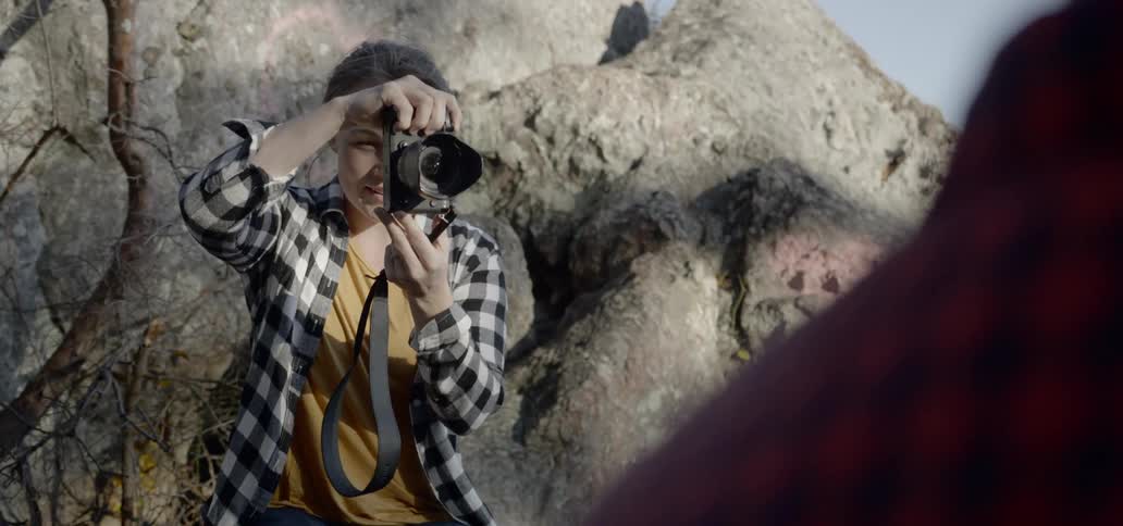 A woman sits outside in front of a large rock formation and raises her camera to take a picture of another person