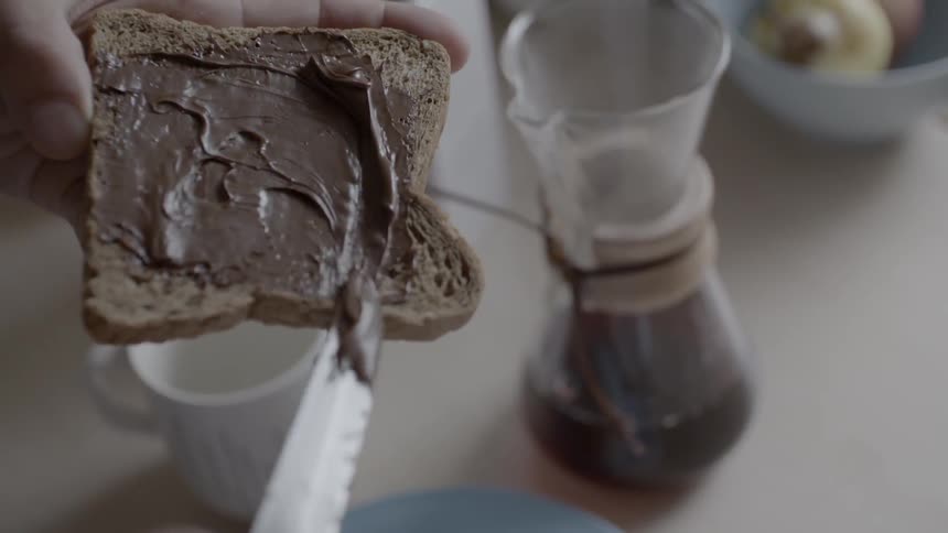 Person spreads hazel butter on spread near brewing coffee
