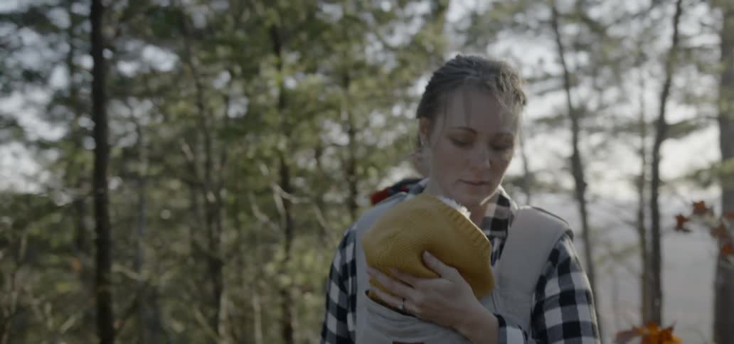 A woman carrying a baby in a front pouch cradles his head as she walks through the woods