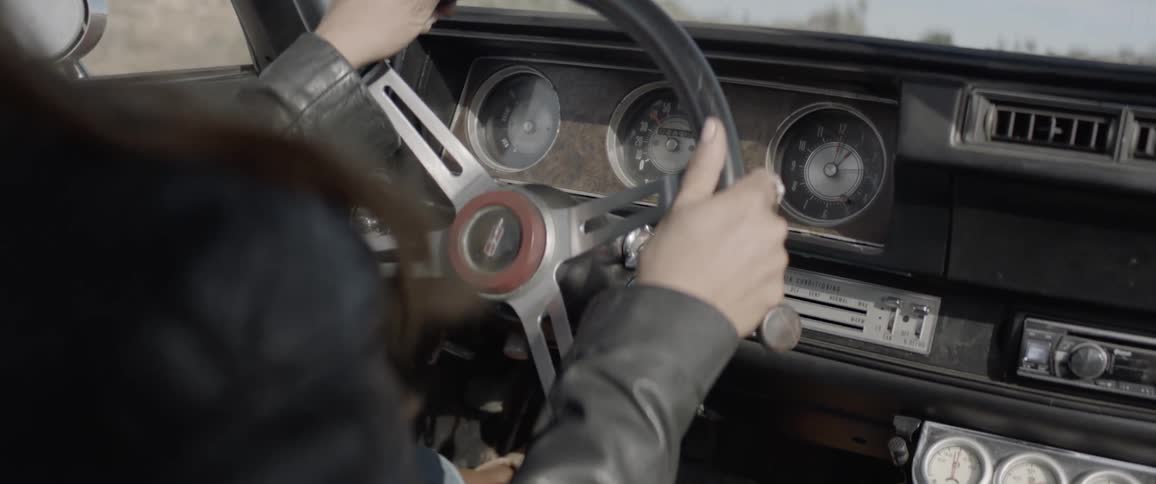A leather jacket wearing person drives a classic car with analogue dials on a sunny day