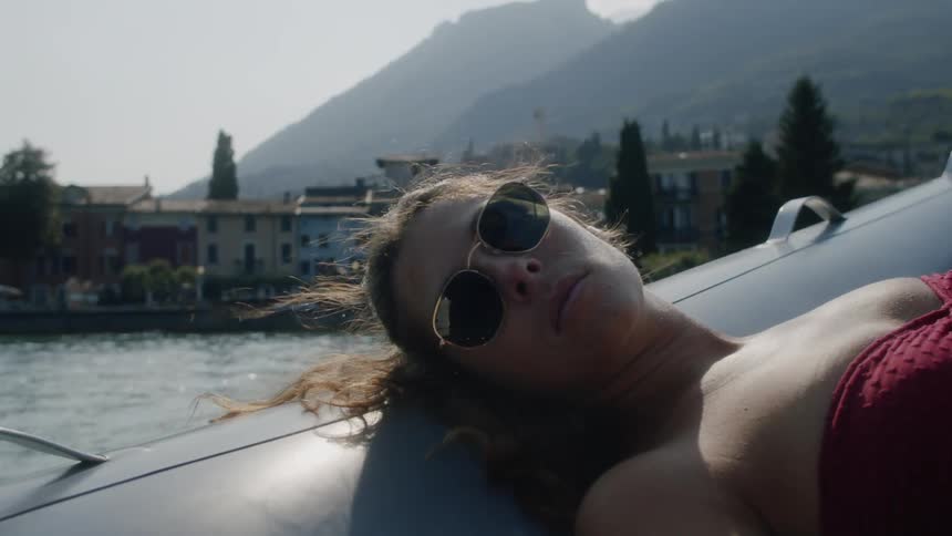 A woman wearing sunglasses relaxes laying her head on the side of a boat which is sailing upstream