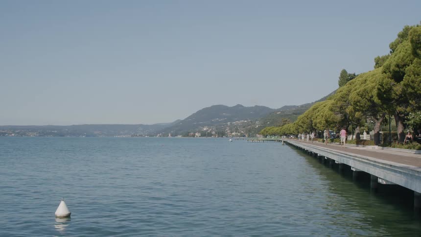 A buoy bobs in the water near a tree lined park path where people are walking