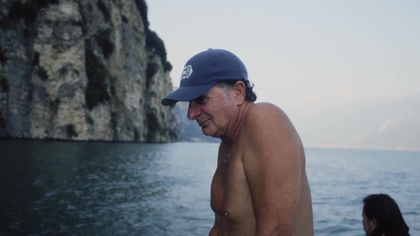 A shirtless man standing up in a boat near the cliffs on shore