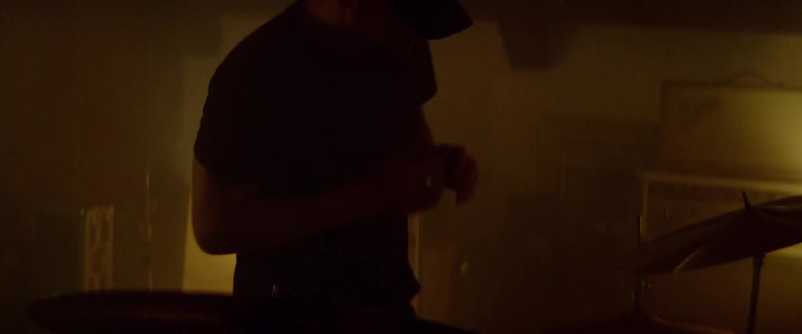 Man wearing cap walks up to drum set and sits down to play in a dark room