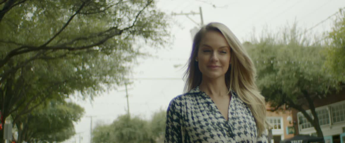 A blonde haired woman smiles as she walks down a street that is lined with trees and shops