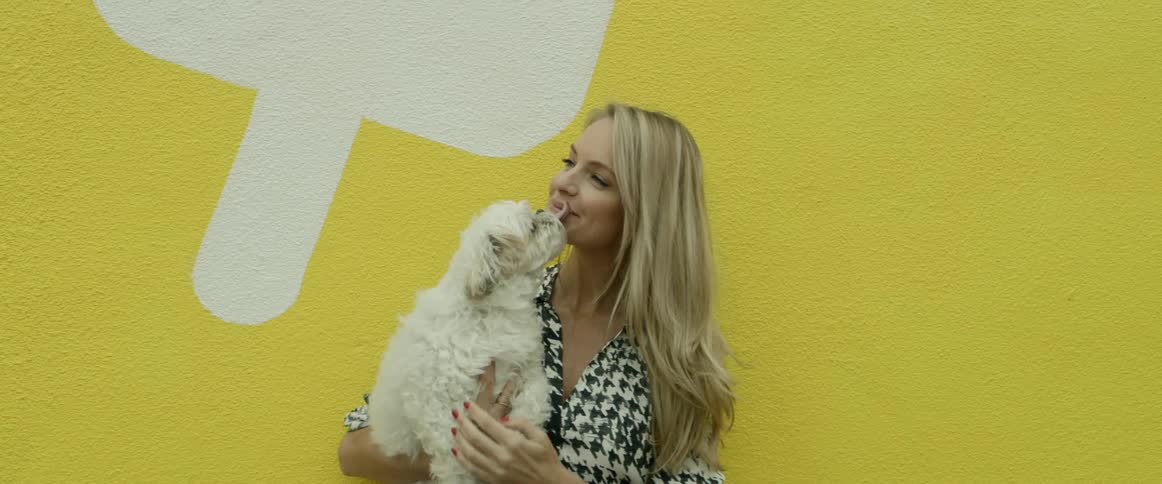 A white female with long blond hair is holding a small white dogand kissing it in front of a yellow wall