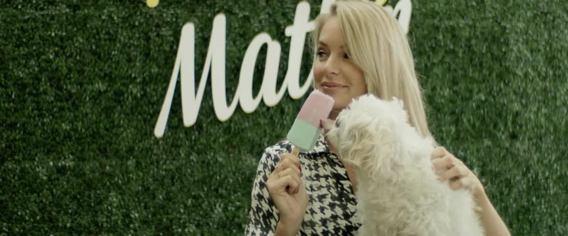 A small white dog licks an ice cream on a stick held by a beautiful blonde woman