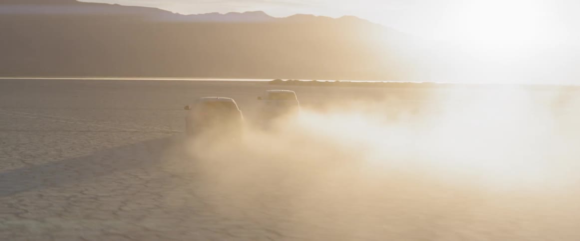 Two vehicles are driving through the desert flats at sunset