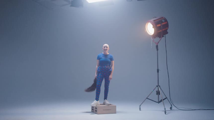 A bald woman stands on set waving her wig around and laughing