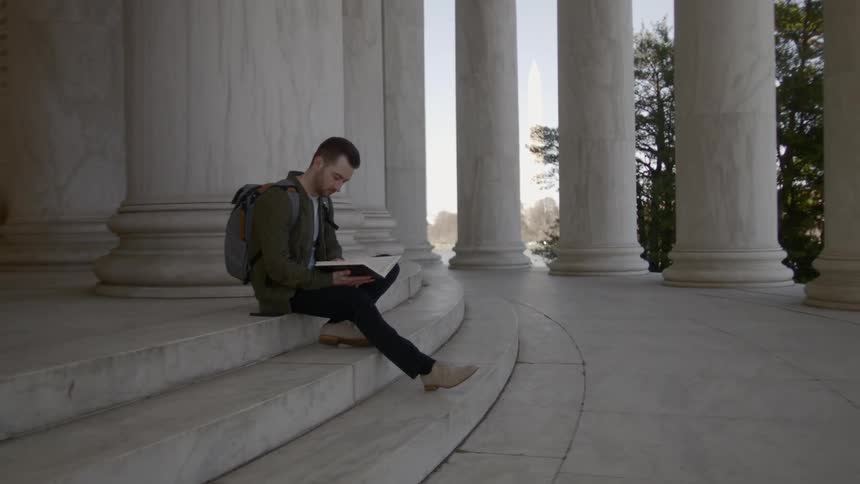 A man sits reading a book on the marble steps of a columned building