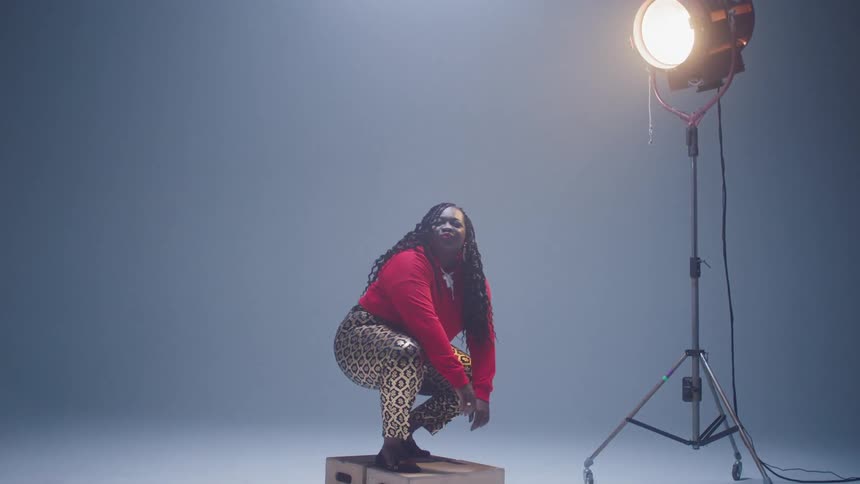 A large black woman dances on a box underneath studio lighting in a studio