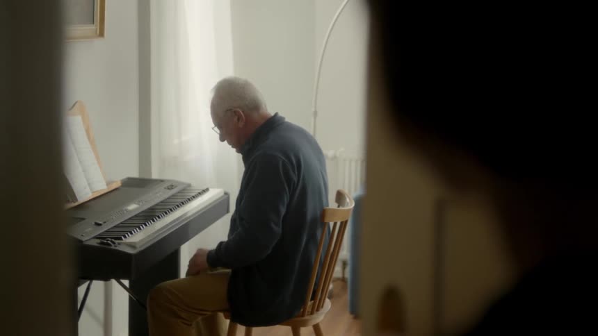 A person watches an old man sitting in a wooden chair play a keyboard