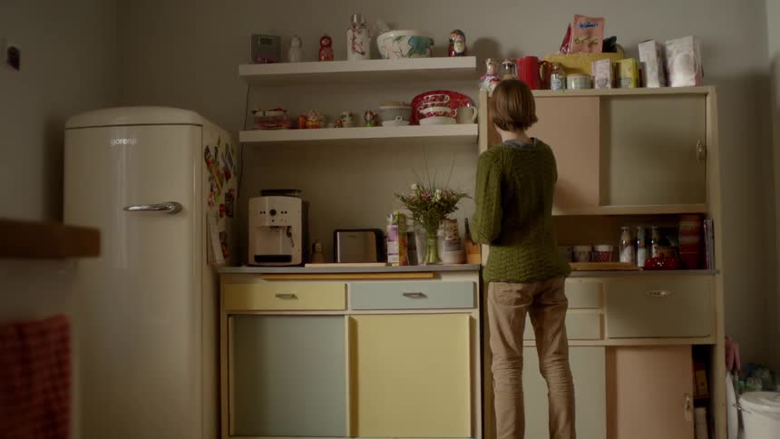 A teenager walks to a kitchen pantry and opens the door to take something out