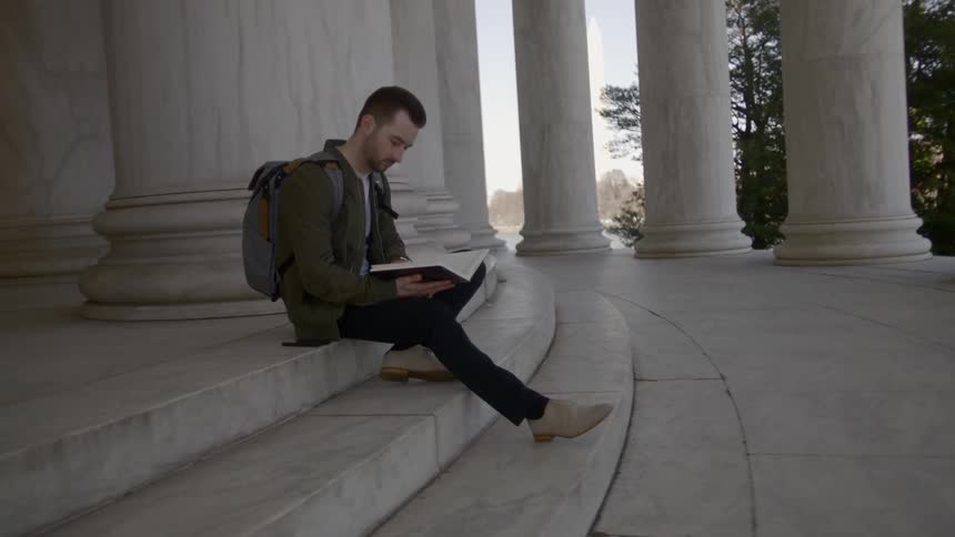 A man sitting on marble steps reading a book with pillars in the background