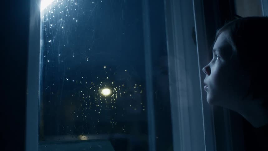 A child is looking out the window at night