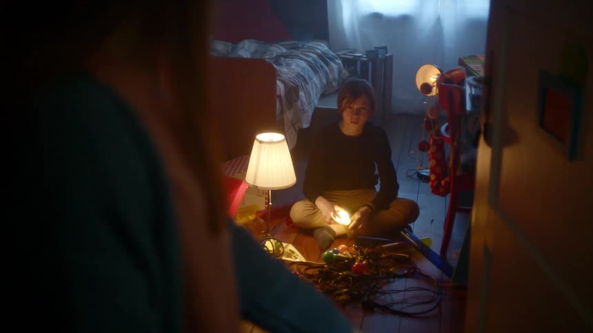 A teenager sits on the ground holding flickering light bulbs as their mom walks past their room