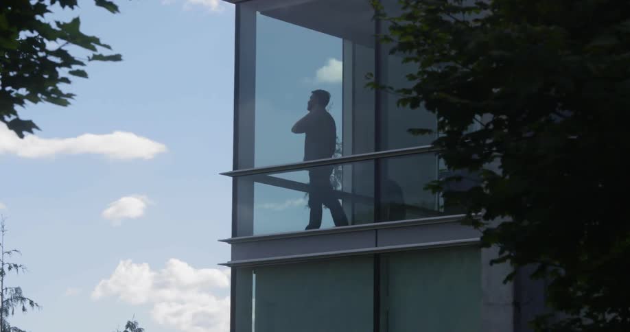 A man talks on the phone and paces back and forth as seen through a window
