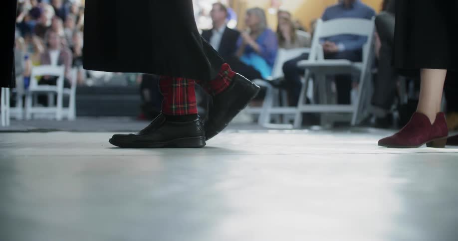 Students feet as they walk in their graduation gown with people sitting on chairs in the background