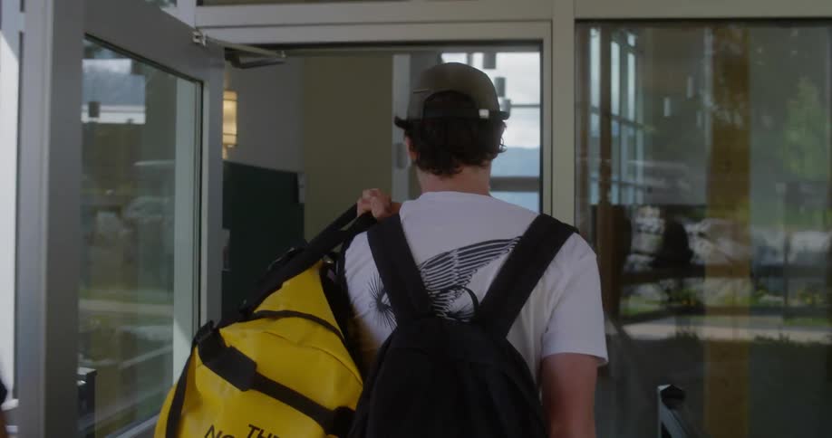 A man holds open the door for another carrying a large yellow duffel and backpack