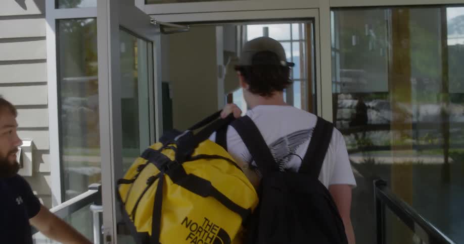 A young man carrying a backpack and large yellow duffel bag is greeted by another person as he enters a building