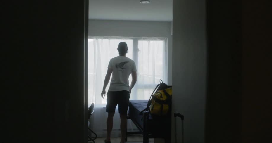 Man pulls the curtains open to look out the window then he sits on a bed