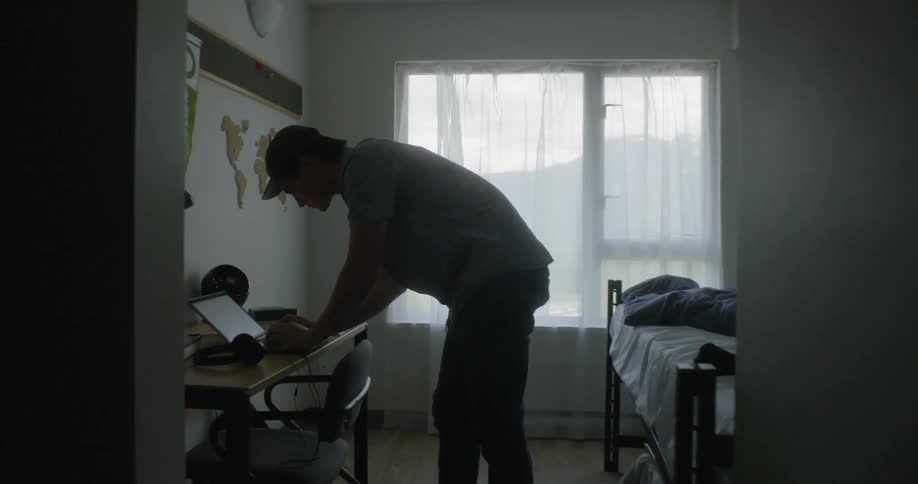 Teenage boy standing over and typing on laptop in his room