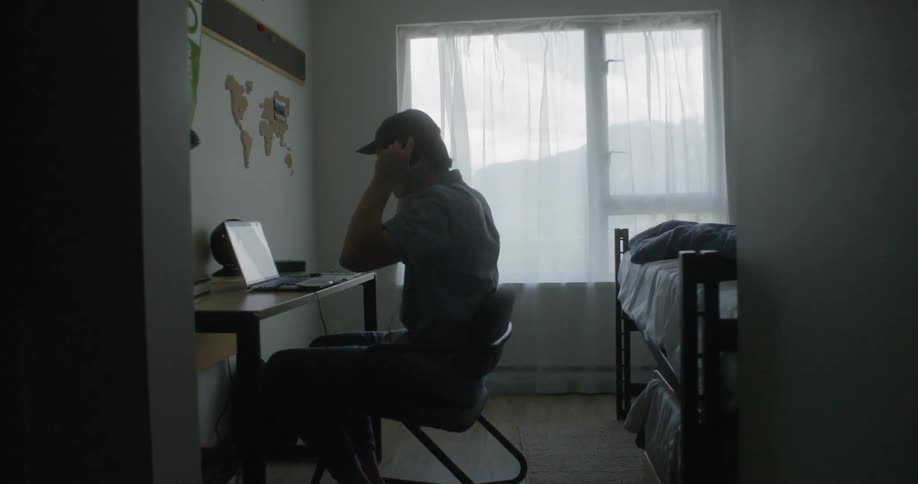 A young man sits at his desk puts his headphones on and then types on his laptop