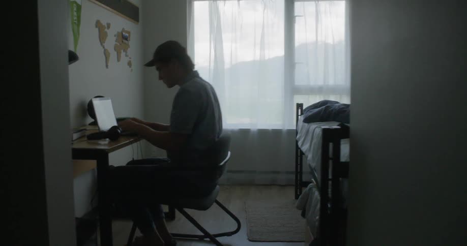 Man wearing a cap sits at the desk to use the laptop and wears the headphones in the room