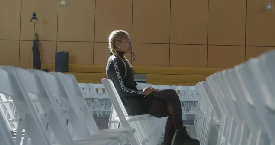 A woman sits alone in a large auditorium filled with white folding chairs