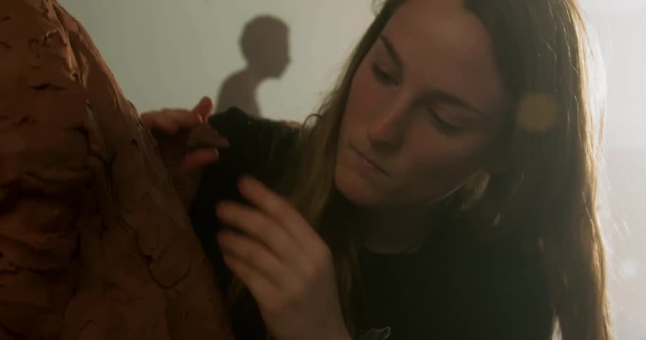 A female takes pieces of clay and adds them to a large clay sculpture before smoothing the surface
