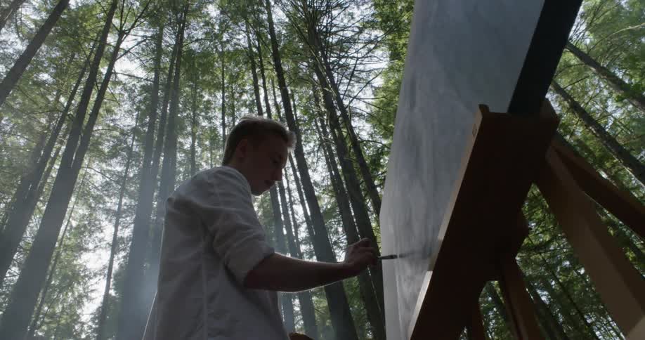 A white male is painting on a canvas on a large painting easelin a forest