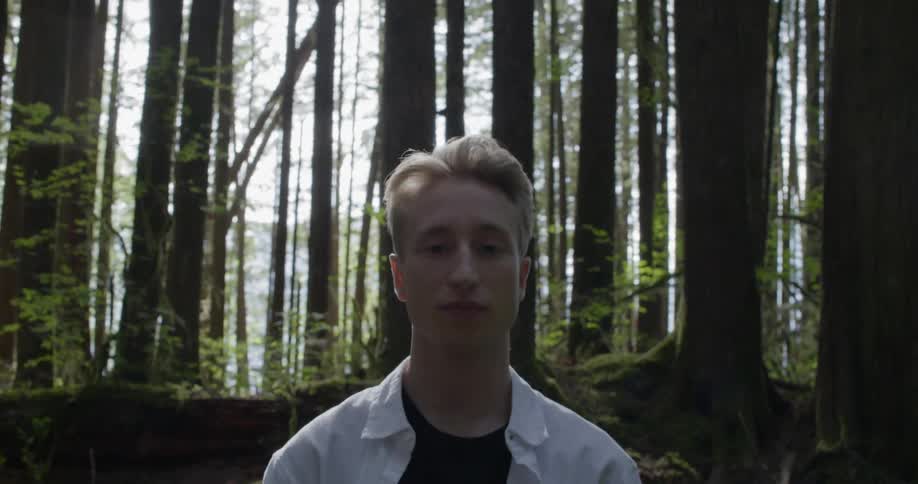 Blonde man wearing a white linen shirt looks at the camera in the dense forest