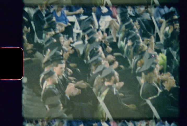 Graduates in caps and gowns applaud as they sit in their seats