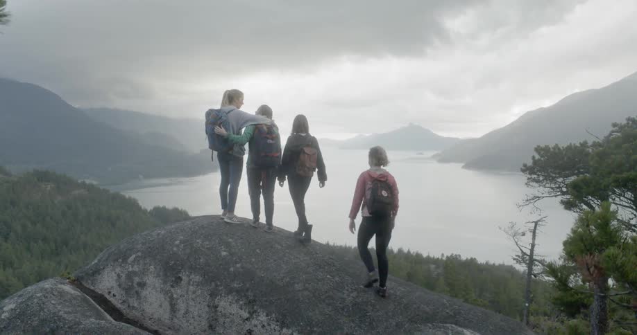 A group of women hike up to the top of a large rock overlooking a lake in a valley