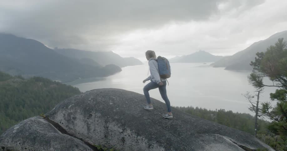 Woman wearing a backpack climbs a hill overlooking a lake under a cloudy sky