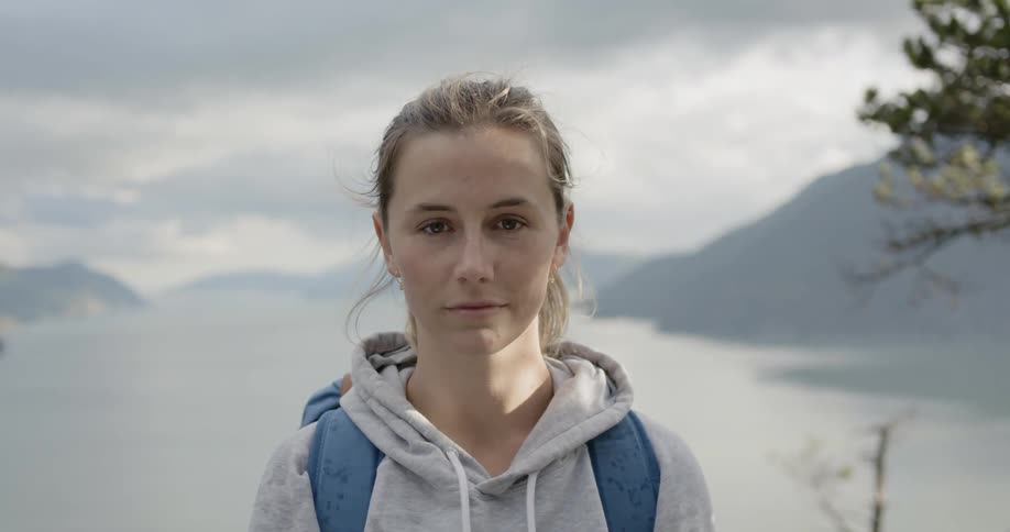 Blonde woman wearing backpack raises her head to look at the camera with the view of the lake and the mountains