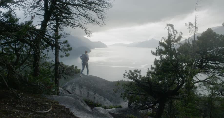 A female hikes up a rock formation and stands at the top of it while looking out at the large body of water below