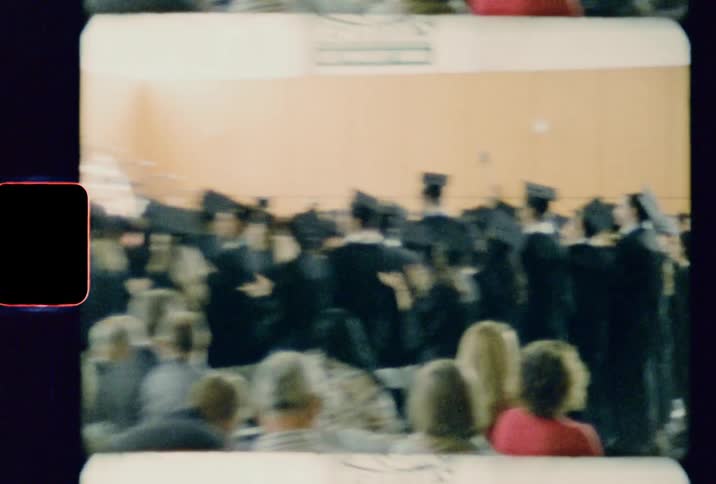 Graduating students standing in a gymnasium clapping while a crowd sits around them
